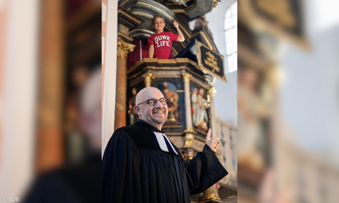 Ein Pfarrer, hinter ihm auf der Kanzel ein Junge im roten Shirt