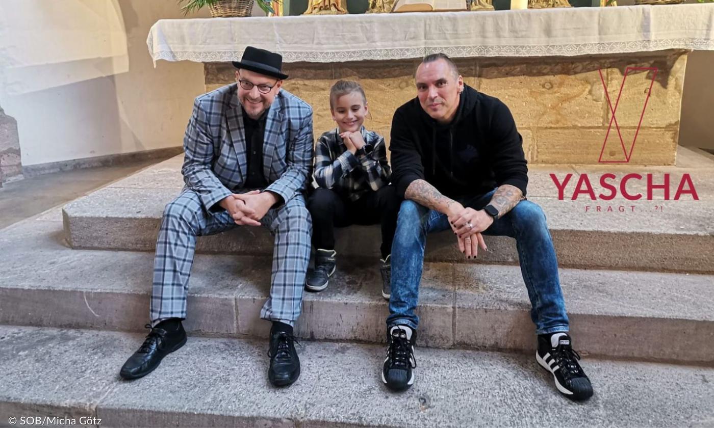 Zwei Männer und ein Junge sitzen vor einem Alter und lachen in die Kamera