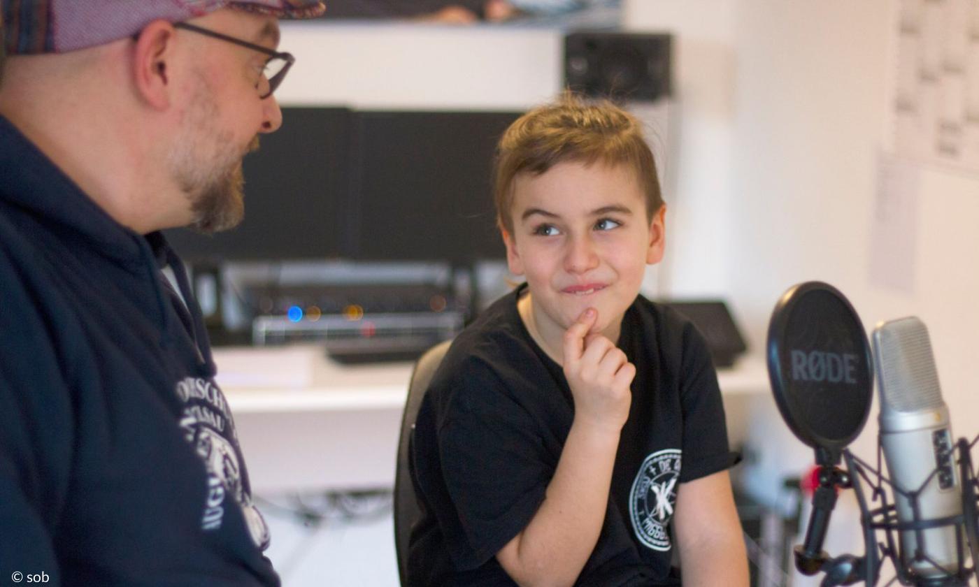 Ein Junge sitzt in einem Aufnahmestudio, vor ihm ein Mikrofon, neben ihm ein Mann