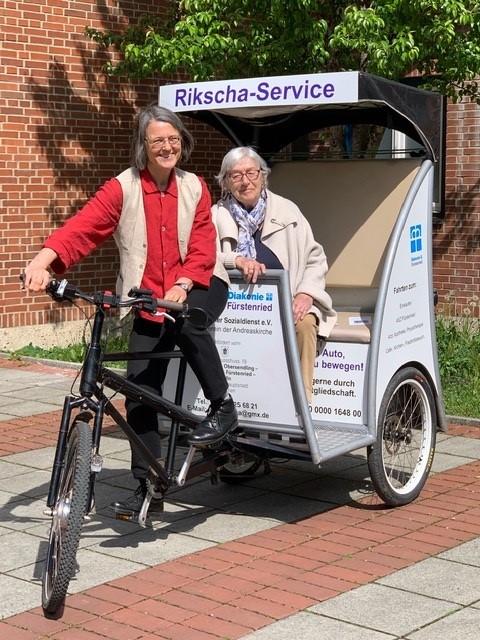 Eine Rikscha mit der Aufschrift "Rikscha-Service", in der eine Seniorin als Fahrgast sitzt. Eine weitere Frau sitzt als Fahrerin vorne auf dem Rad.