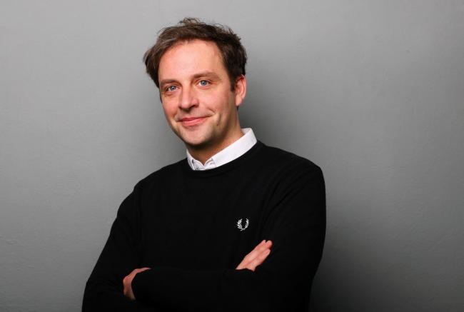 Nils Kumkar vor einer grauen Wand, die Arme verschränkt, in die Kamera lächelnd