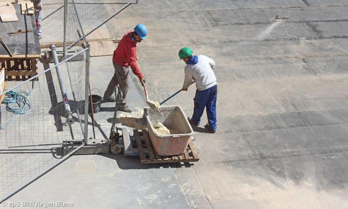 Zwei Arbeiter mit blauem und grünem Helm schaufeln Sand in einen Kübel auf einer Baustelle. Links neben ihnen sieht man einen Bauzaun