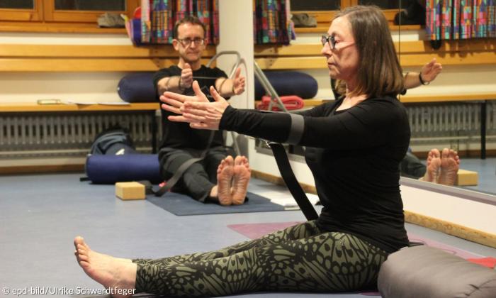In einem Gymnastikraum mit Spiegelwand rechts im Bild sitzt eine Frau mit ausgestreckten Beinen auf dem Boden. Ihre Arme häält sie ausgestreckt nach vorne. Im Hintergrund sitzt ein Mann an eine Bank angelehnt in der gleichen Position.