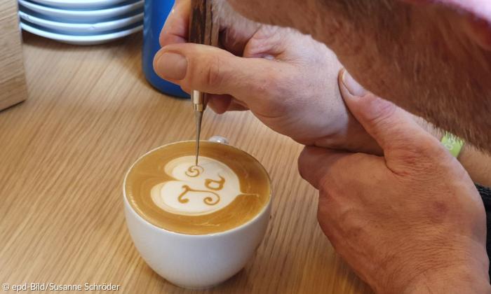 Ein Mann graviert in den Schaum eines Kaffees mit einer Art Pinsel das Wort Ja. Das Bild zeigt ihn von oben herab. Der Kaffee steht in der Mitte.