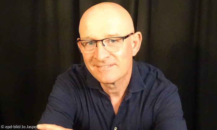 Portrait eines Mannes mit Glatze und Brille. Er trägt ein blaues Langarmhemd.