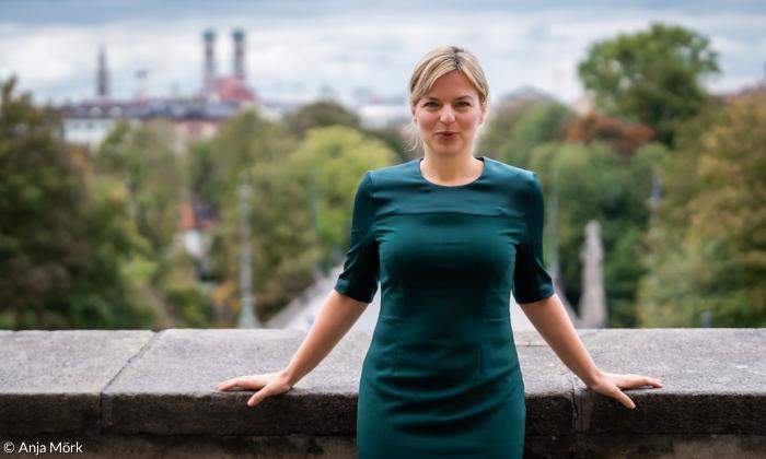 Eine blonde Frau im grünen Kleid, dahinter die Türme der Münchner Frauenkirche