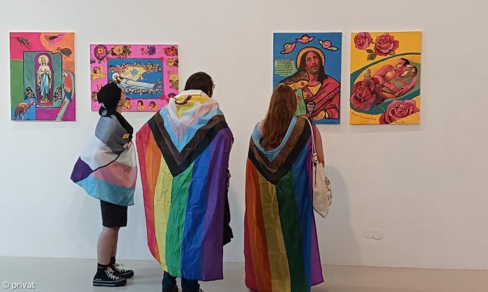 Drei Personen, die mit dem Rücken zum Betrachter stehen, schauen sich gerade bunte Gemälde an. Sie tragen Fahnen der LGBTQIA*-Bewegung.