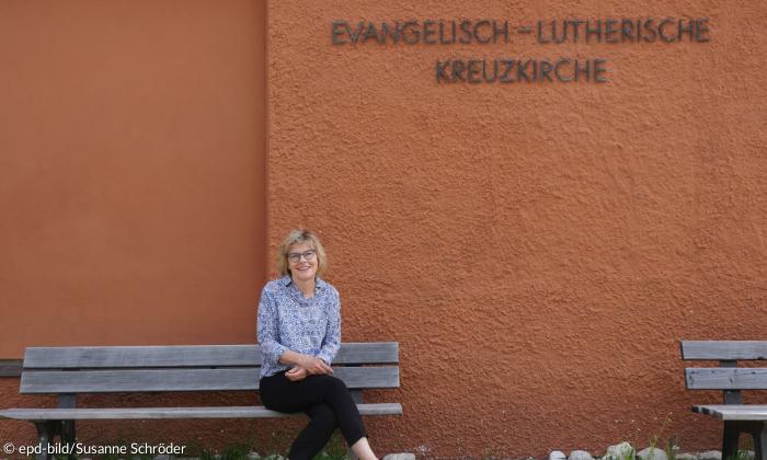 Eine Frau sitzt auf einer Bank vor einer rotbraunen Wand, auf der "Evangelisch-Lutherische Kreuzkirche" steht