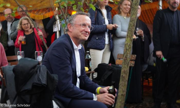 Ein Mann im Anzug, sitzend, er lächelt verschmitzt