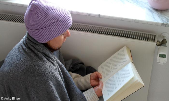 Eine Frau sitzt mit Mütze und Decke an der Heizung