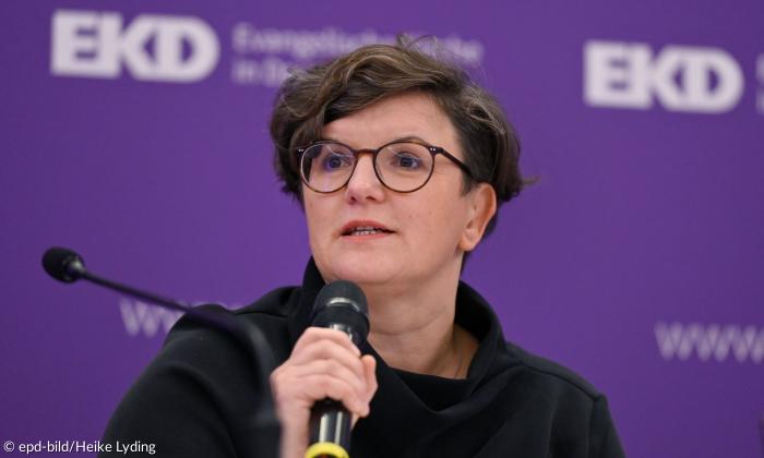 Eine Frau mit einem Mikrofon, hinter ihr der Schriftzug "EKD" auf lila Hintergrund