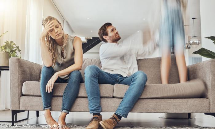 Eine Frau greift sich an den Kopf, neben ihr auf der Couch sitzt ein Mann, der nach einem Kind greift, das auf der Couch steht