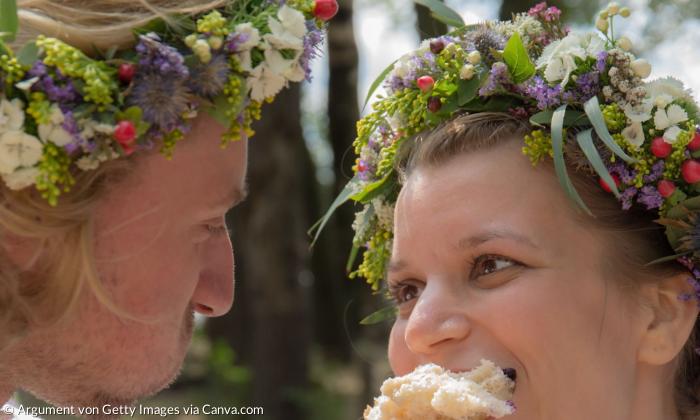 Ein Hochzeitspaar mit Blumenkränzen. Sie schauen sich an und essen eine Semmel.