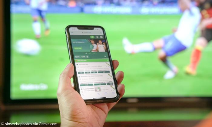 Ein Handy, auf dem Wetten zu sehen sind, dahinter ein Fernseher, auf dem Fußball läuft