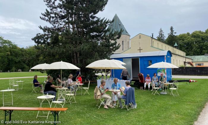 Vor einer Kapelle steht ein Bauwagen auf einer Wiese, davor Tische mit Schirmchen, an denen Menschen sitzen und Kaffee trinken