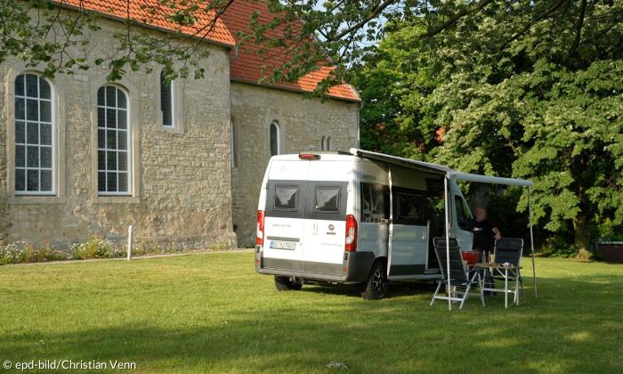 Vor einer Kirche steht ein Wohnmobil auf der grünen Wiese, davor steht ein Mann