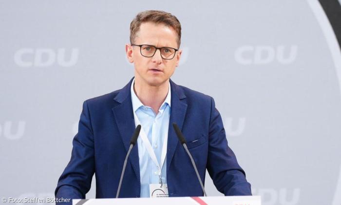 Ein Mann mit dunkelblauer Anzugjacke und Brille spricht in ein Mikrofon