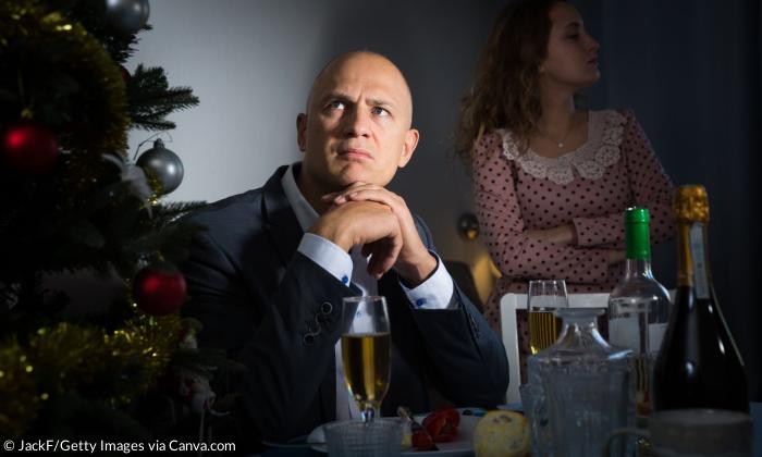 Ein Mann sitzt am Tisch und schaut genervt, daneben ein Weihnachtsbaum, im Hintergrund eine Frau, die die Arme verschränkt und in die andere Richtung schaut