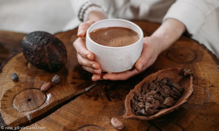 Zwei Hände halten eine Tasse Kakao, drumherum sind Kakaobohnen zu sehen