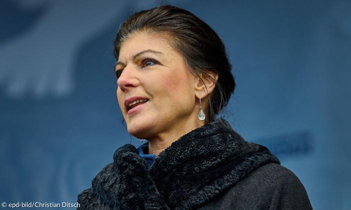 Eine Frau vor einer blauen Wand, sie trägt einen schwarzen Mantel mit auffälligem Kragen