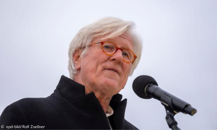Ein Mann mit weißen Haaren und rötlicher Brille spricht in ein Mikrofon