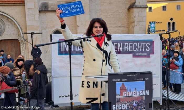 Eine Frau steht auf einer Bühne, umringt von Menschen. Sie hält ein Schild hoch: "Kein Platz für Nazis"
