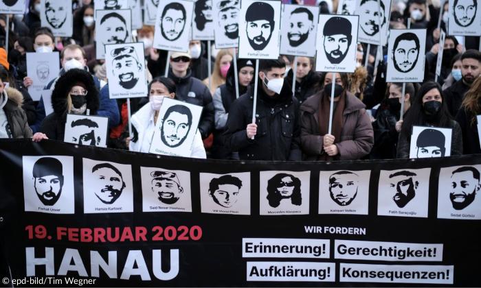 Menschen vor einem Transparent, auf dem acht Gesichter zu sehen sind, sie halten Schilder hoch, auf denen auch Gesichter zu sehen sind