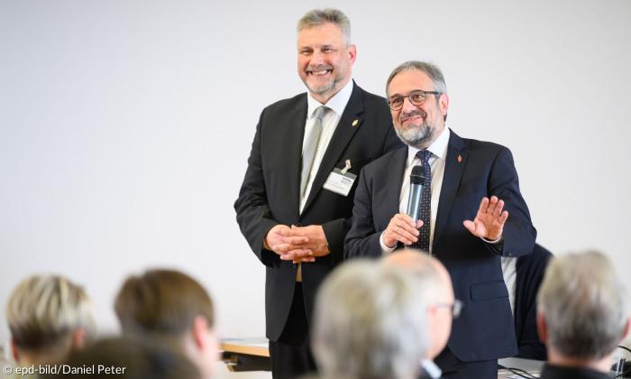 Zwei Männer in dunklen Anzügen stehen auf einer Bühne vor Leuten