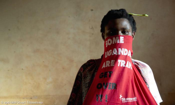 Eine Frau hat eine rote Maske vor der unteren Gesichtshälfte, auf der "Some Ugandans are trans get over it" steht