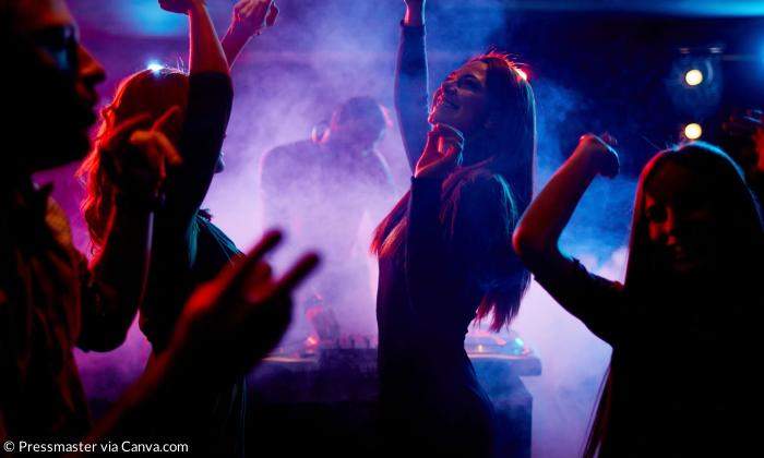 Menschen tanzen im Discolicht, im Hintergrund ein DJ