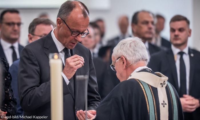 Ein Mann steht vor einem Priester mit gesenktem Kopf, dahinter Männer in Anzügen