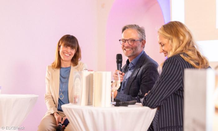 Drei Menschen sitzen an einem Tisch, ein Mann in der Mitte hält ein Mikrofon
