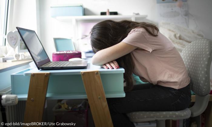 Ein Mädchen sitzt am Tisch und hat den Kopf in den Armen vergraben, vor ihr ein aufgeklapptes Laptop
