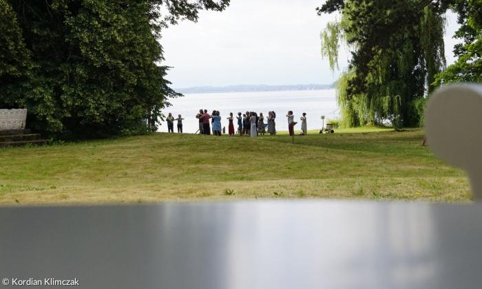Teilnehmende stehen in einem Kreis auf einer Wiese am See, Bewegung oder Achtsamkeit verbunden mit der Natur.