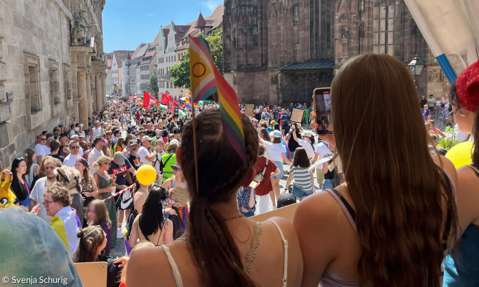 Zwei Mädchen von hinten mit Regenbogenflagge vor einer Menschenmenge