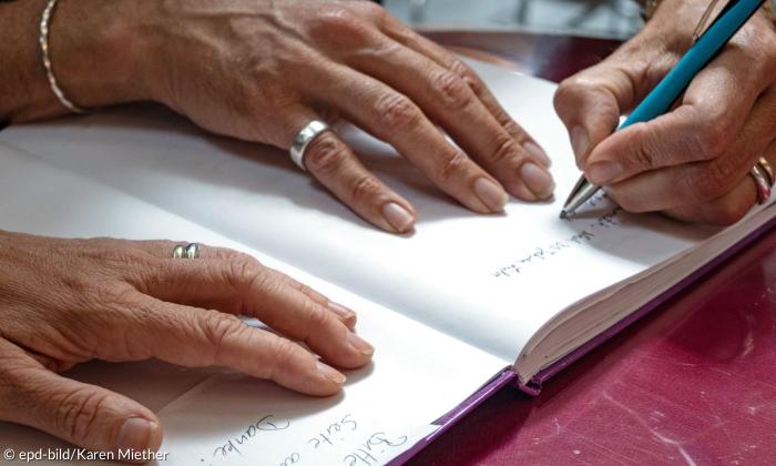 Eine Hand schreibt in ein Buch, zwei weitere liegen auf den Seiten