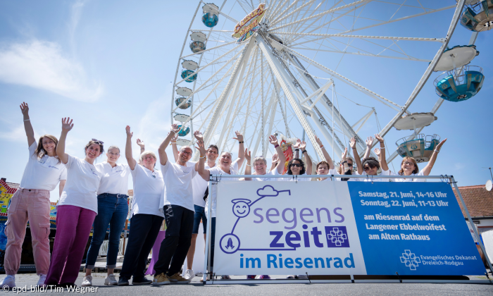 Auf dem Bild sieht man Menschen vor einem Riesenrad. Davor ist ein Banner mit der Aufschrift "Segenszeit im Riesenrad". 