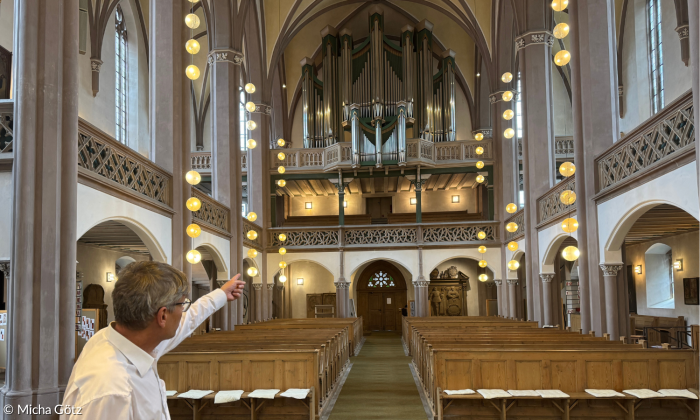 Pfarrer Ulrich Winkler in der Petrikirche Kulmbach mit der imposanten Orgel im Hintergrund, auf die der Pfarrer mit der Hand zeigt.