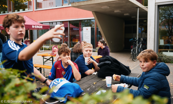 Eine Gruppe von Jugendlichen sitzt draußen auf Bänken, isst Snacks, trinkt Getränke und unterhält sich angeregt.