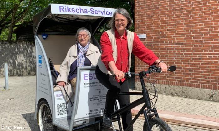 Eine Rikscha mit der Aufschrift "Rikscha-Service" und einer älteren Frau, die als Fahrgast darin sitzt und einer Frau, die als Fahrerin vorne auf dem Fahrrad sitzt