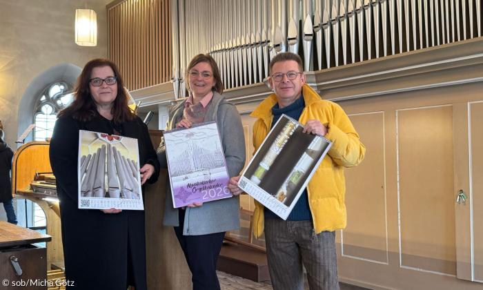 Drei Menschen zeigen Kalender vor einer Orgel