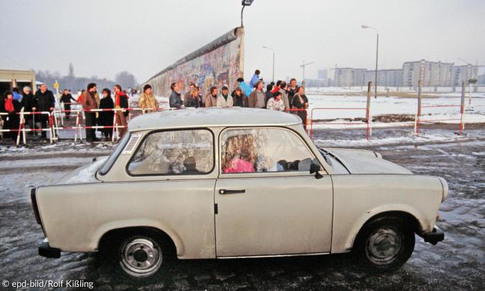 Ein Trabant fährt auf der Straße, dahinter Reste der Berliner Mauer