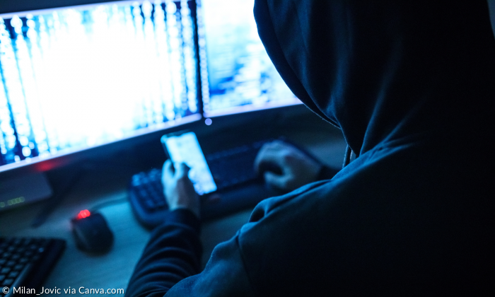 Person mit Hoodie über Kopf von hinten in einem dunklen Raum vor Handy und PC.