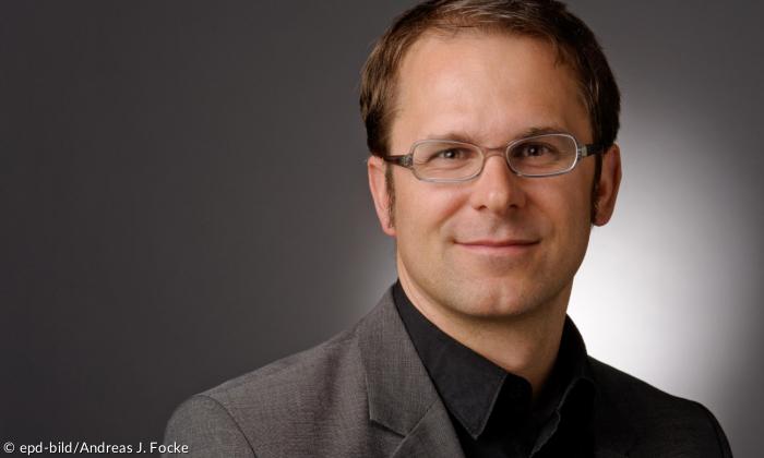 Ein Mann mit Brille, der in die Kamera lächelt. Grauer Hintergrund.