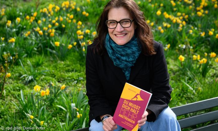 Melanie Dertinger hält ihr Buch in der Hand und kniet vor einem blühenden Wiese