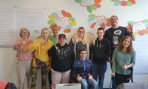 Neun Menschen stehen oder sitzen vor einer Wand mit bunten Aufklebern und heben ihren Daumen