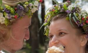 Ein Hochzeitspaar mit Blumenkränzen. Sie schauen sich an und essen eine Semmel.