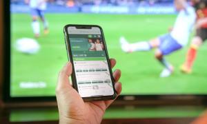 Ein Handy, auf dem Wetten zu sehen sind, dahinter ein Fernseher, auf dem Fußball läuft