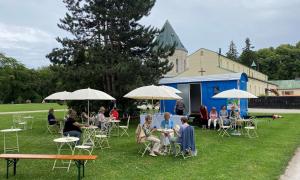 Vor einer Kapelle steht ein Bauwagen auf einer Wiese, davor Tische mit Schirmchen, an denen Menschen sitzen und Kaffee trinken