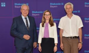 Zwei Männer und eine Frau stehen vor einer lila Wand mit der Aufschrift "Diakonie Bayern"